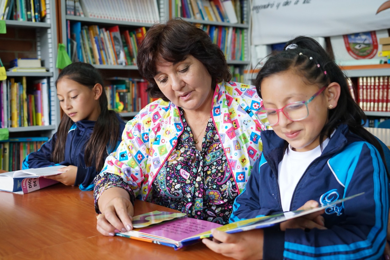 Maestra en clase con dos estudiantes leyendo un libro