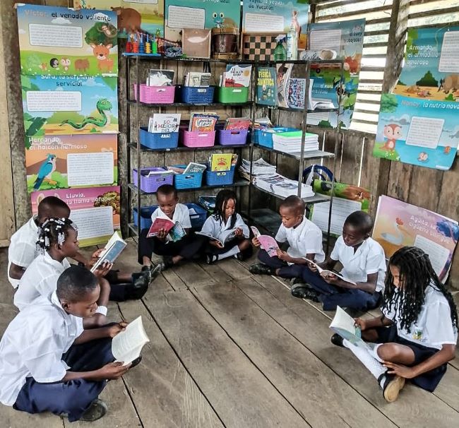 Imagen de niños estudiantes sentados en el piso, dentro del aula de clase, leyendo cada uno un libro
